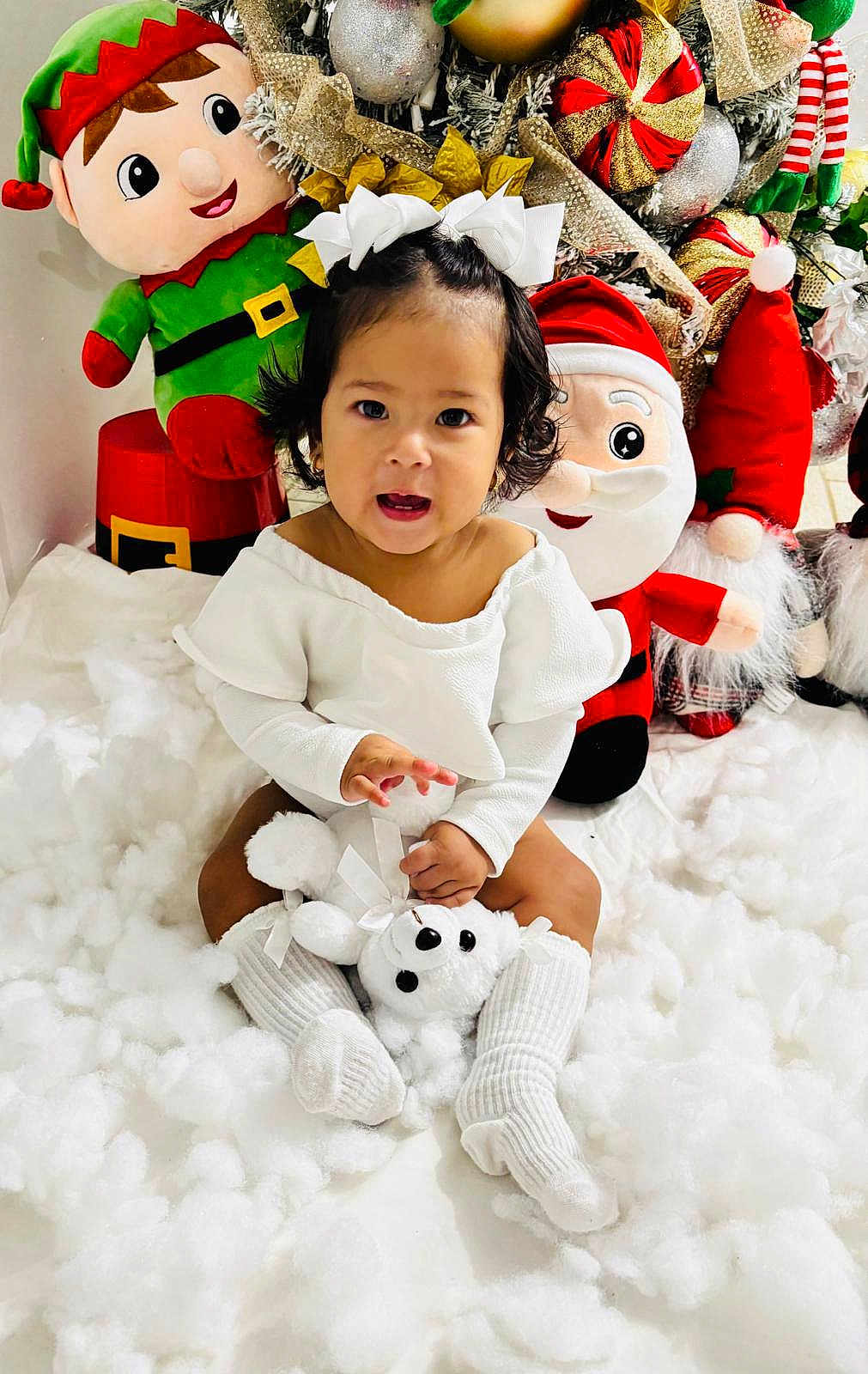 Eva Mota is registered to the contest to win money with this photo: toddler, child, white_clothing, white_socks, teddy_bear, christmas_decor, plush_toys, elf_toy, santa_toy, festive, holiday, snow_effect, indoor, cute, smiling, sitting, holiday_season, decorations, toy, happy