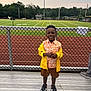 child, boy, smiling, standing, bleachers, chain_link_fence, sports_field, running_track, stadium_lights, trees, grass, outdoor, casual_clothing, jacket, shorts, sneakers, daytime, happy, person, background