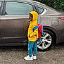 child, raincoat, yellow, backpack, car, rain, wet, pavement, sneakers, smile, hood, blue_pants, vehicle, outdoor, person, side_view, tree, greenery, urban, weather