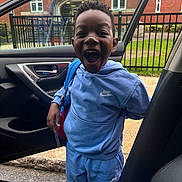 Joash is registered to the contest to win money with this photo: child, boy, blue_clothing, nike, tracksuit, backpack, car_door, open_car_door, urban, brick_building, fence, excited, happy, standing, outdoor, smiling, person, daylight, road, sidewalk