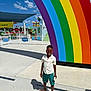 child, boy, sunglasses, rainbow, amusement_park, outdoor, summer, sunny, shorts, shirt, flip_flops, smiling, shadow, concrete, fence, clouds, blue_sky, playground, colorful, fun