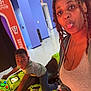woman, child, arcade_game, motorcycle, indoor, playful, tongue_out, casual_clothing, trampoline_park, blue_wall, orange_sign, game_console, joyful, fun, braids, bracelets, jeans, grey_tank_top, black_hair, floor
