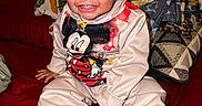 Miloh is registered to the contest to win money with this photo: toddler, child, smiling, mickey_mouse, hoodie, sitting, bed, pillow, blanket, indoor, top_bun, happy, cute, clothing, person, portrait, young_child, cozy, room, casual