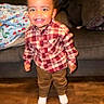 toddler, child, smiling, plaid_shirt, brown_pants, white_shoes, indoor, couch, blanket, wooden_floor, happy, standing, person, face, home, cozy, warm, cute, portrait, clothing