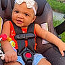baby, bow_headband, bracelet, brick_wall, car_seat, child, close_up, denim_shorts, earring, greenery, happy, infant, outdoor, photography, portrait, red_outfit, safety_harness, sitting, smiling, toy