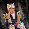 baby, child, infant, smiling, car_seat, seatbelt, bow, floral_clothing, pink_shirt, patterned_pants, portrait, sitting, indoors, vehicle, safety_harness, fashion, hand_to_mouth, happy, closeup, photography