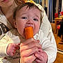 baby, child, carrot, eating, hand, adult, indoor, person, holding, face, clothing, white, skin, hair, table, chair, curtain, window, floor, warm_light