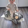 toddler, child, toy_car, indoor, rug, wooden_floor, sweater, pants, plush_toy, steering_wheel, modern_room, curly_hair, cute, playtime, flooring, furniture, minimalistic, light, smiling, person