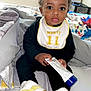 Kamari is registered to the contest to win money with this photo: baby, child, infant, bib, sneakers, bed, blanket, tube, cream, window, vertical_blinds, curly_hair, topknot, indoors, person, clothing, footwear, sitting, young_child, cute