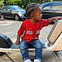 toddler, child, red_sox, jersey, jeans, sneakers, scooter, outdoor, side_view, street, urban, casual, parked_cars, greenery, daylight, person, fashion, curly_hair, playful, young