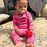 baby, child, toddler, sitting, indoors, living_room, carpet, couch, pink_pajamas, pacifier, toy, red_toy, curly_hair, portrait, face, eyes, earring, barefoot, cozy, home