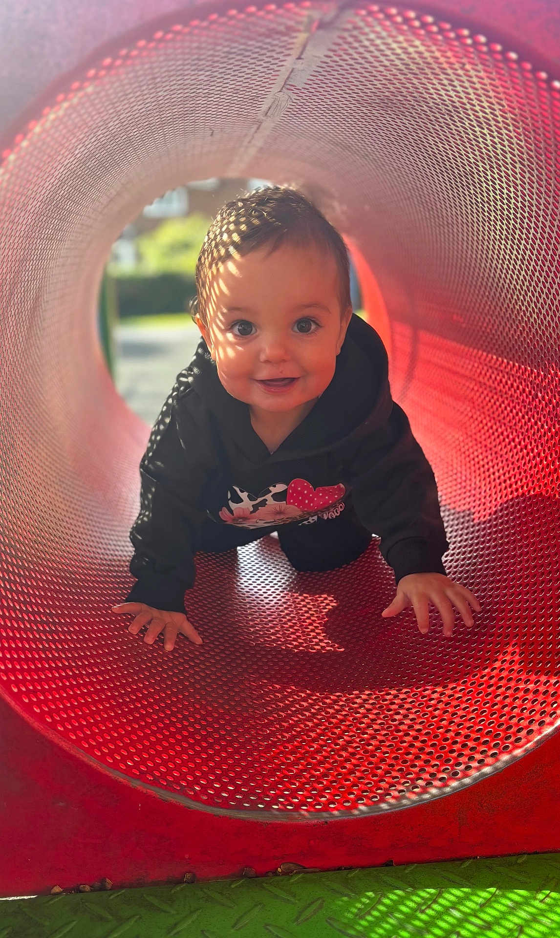 Ella is registered to the contest to win money with this photo: child, toddler, crawling, playground, red_tunnel, sunlight, shadow_pattern, happy, hoodie, outdoor, daylight, smile, face, curious, play, metal, perforated_surface, cute, young_child, fun
