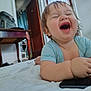 Keith is registered to the contest to win money with this photo: baby, laughing, indoor, bed, light_blue_shirt, phone, yellow_object, wooden_table, wicker_chair, furniture, happy, child, smile, person, curly_hair, plush, home, cozy, young_child, playful