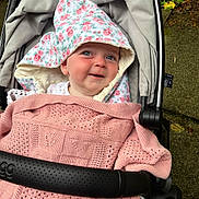 Maggie is registered to the contest to win money with this photo: baby, child, stroller, blanket, pink, knitted, hooded_towel, floral_pattern, smiling, face, blue_eyes, outdoor, pavement, greenery, fabric, cozy, cute, person, infant, portrait