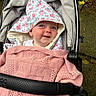 baby, child, stroller, blanket, pink, knitted, hooded_towel, floral_pattern, smiling, face, blue_eyes, outdoor, pavement, greenery, fabric, cozy, cute, person, infant, portrait