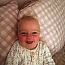 Maggie is registered to the contest to win money with this photo: baby, smiling, face, child, pillow, bed, pink, checkered, clothing, floral, happy, infant, cute, rosy_cheeks, portrait, lying_down, indoors, person, soft_lighting, cozy