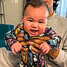 Koa joined the competition — help win amazing prizes! baby, child, infant, smiling, happy, portrait, indoor, caregiver, hands, holding, colorful_clothing, patterned_outfit, open_mouth, blue_eyes, short_hair, seated, couch, home, close_up, family