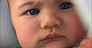 Koa is registered to the contest to win money with this photo: baby, infant, child, face, portrait, close_up, eyes, serious, green_clothing, outfit, fist, hand, indoor, blanket, cuddled, head, skin, photography, baby_portrait, looking_at_camera