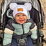 baby, child, stroller, jacket, hat, pom_poms, smile, happy, outdoor, pavement, baby_clothing, winter_wear, cute, infant, person, seat_belt, footwear, cold_weather, baby_stroller, urban