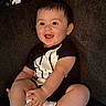 baby, child, smiling, happy, sitting, couch, clothing, black_shirt, white_shoes, indoor, person, infant, cute, portrait, young_child, hands, feet, face, hair, comfort