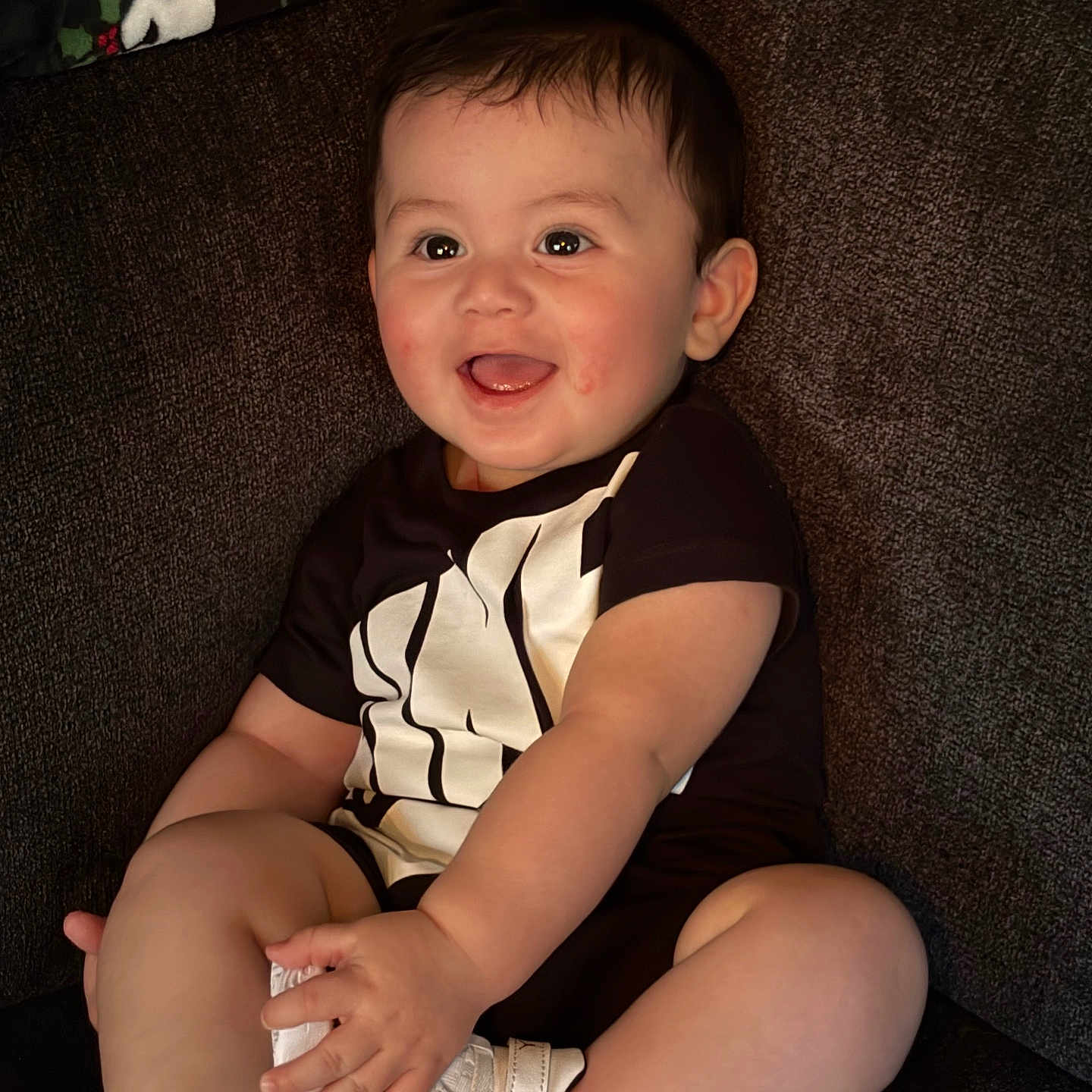 Tommy joined the competition — help win amazing prizes! baby, child, smiling, sitting, happy, couch, clothing, white_sneakers, black_shirt, person, indoor, portrait, skin, face, short_hair, cute, infant, small_hands, legs, footwear