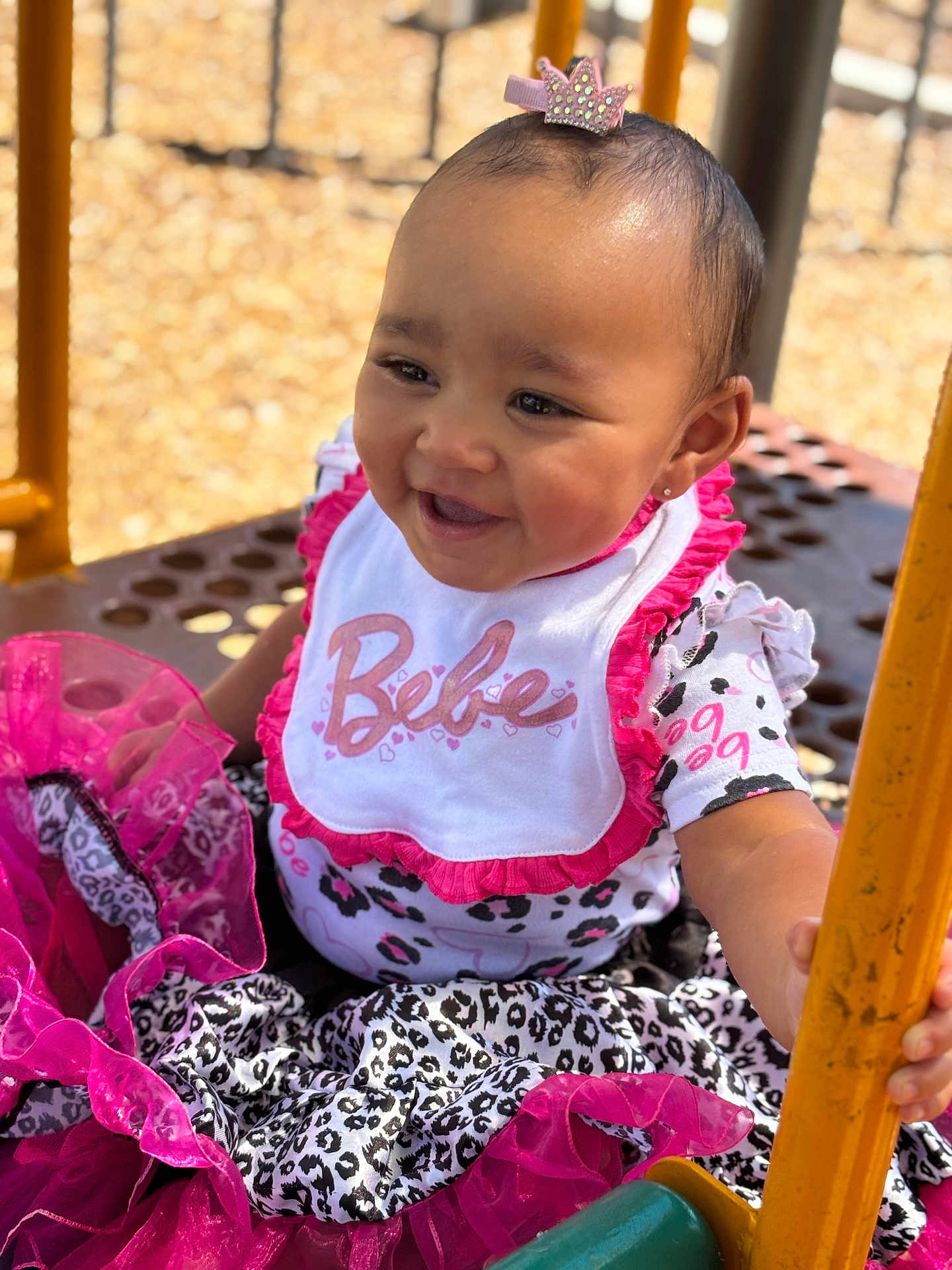 Genesis is registered to the contest to win money with this photo: baby, child, smiling, pink, bib, crown, hair_clip, leopard_print, playground, outdoor, sunny, happy, dress, clothing, hand, yellow_bar, sitting, person, cute, infant