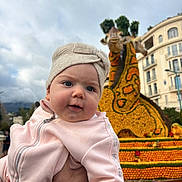 Jodie participe au concours pour gagner de l'argent avec cette photo : baby, child, infant, pink_clothing, headwrap, hand, outdoor, flower_sculpture, giraffe, statue, cloudy_sky, building, urban, person, portrait, cute, floral_art, daytime, nature, celebration