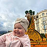 Jodie participe au concours pour gagner de l'argent avec cette photo : baby, child, smiling, face, pink_clothing, headwear, turban, giraffe_sculpture, fruit_art, orange_fruit, outdoor, daytime, sky, cloudy, building, urban, festival, happy, portrait, cute