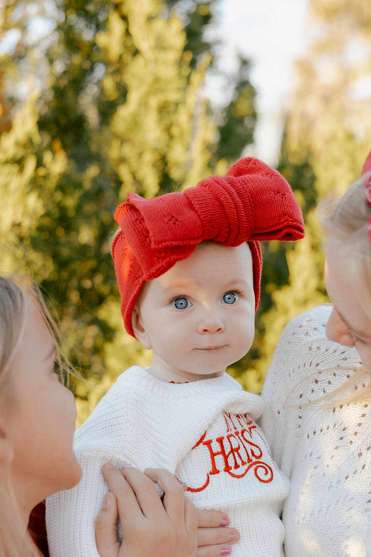 Alexis joined the competition — help win amazing prizes! baby, child, red_bow, hat, white_sweater, adult_hands, outdoor, tree, greenery, portrait, blue_eyes, cute, holiday, family, winter_clothing, knitwear, soft_light, holding, closeup, smiling