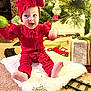 Alexis is registered to the contest to win money with this photo: baby, child, red_clothing, head_wrap, christmas_tree, ornament, holiday_decor, carpet, toy_train_track, gift_box, smiling, sitting, indoor, festive, holiday, joyful, cute, happy, decorations, celebration