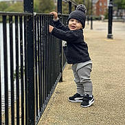 Elias is registered to the contest to win money with this photo: toddler, child, boy, standing, winter_clothing, hat, sweater, pants, sneakers, outdoor, railing, pavement, urban, daytime, smiling, face, person, nature, background, playful
