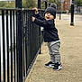 toddler, child, boy, standing, winter_clothing, hat, sweater, pants, sneakers, outdoor, railing, pavement, urban, daytime, smiling, face, person, nature, background, playful
