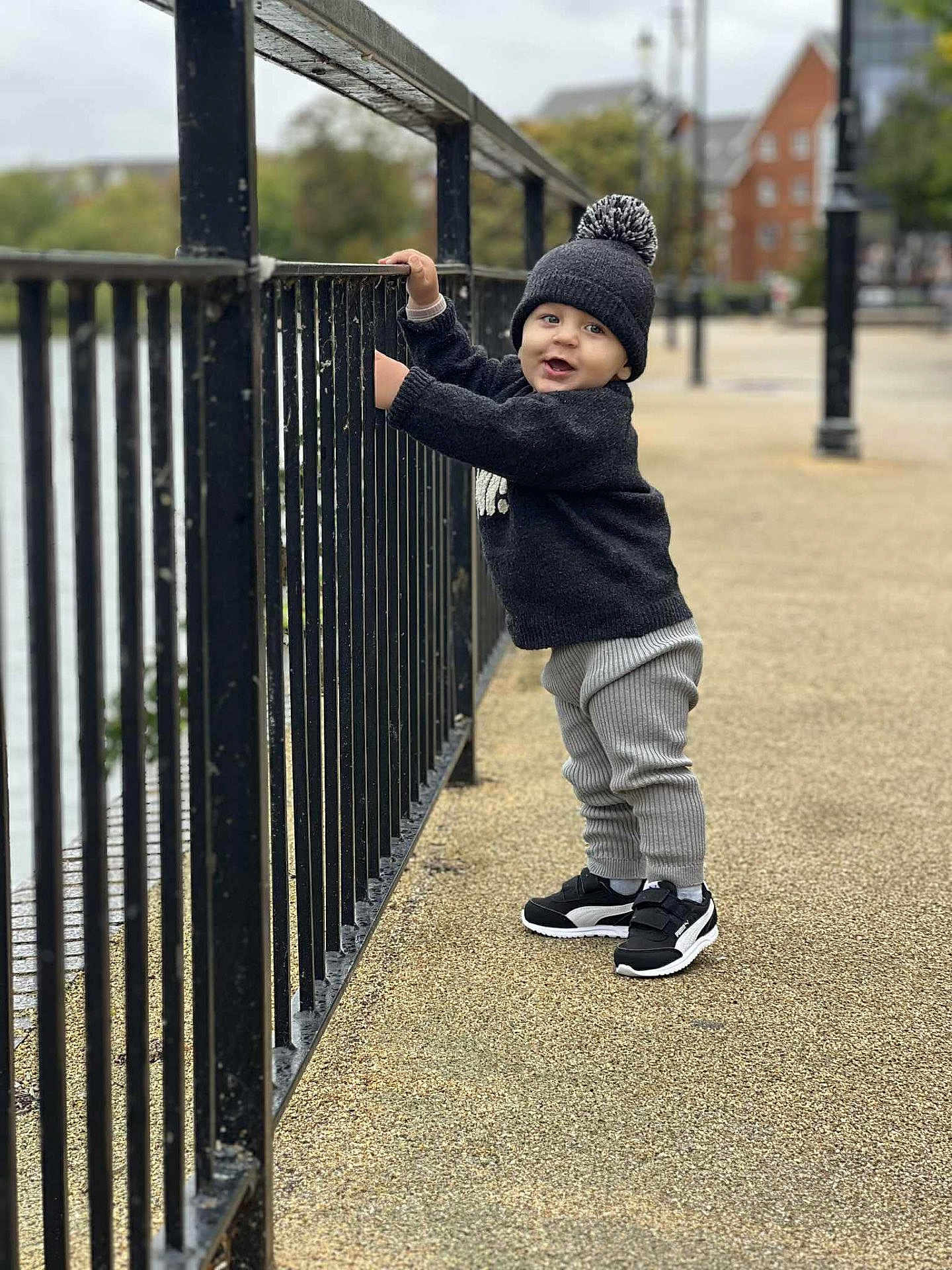 Elias is registered to the contest to win money with this photo: toddler, child, boy, standing, winter_clothing, hat, sweater, pants, sneakers, outdoor, railing, pavement, urban, daytime, smiling, face, person, nature, background, playful