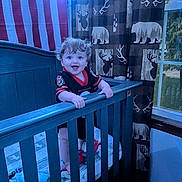 Parker is registered to the contest to win money with this photo: toddler, crib, child, curly_hair, smiling, indoor, curtains, window, bedroom, blue_crib, baby, clothing, patterned_curtains, toy, happy, person, wooden_furniture, standing, home, football_jersey