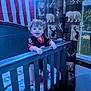 toddler, crib, child, curly_hair, smiling, indoor, curtains, window, bedroom, blue_crib, baby, clothing, patterned_curtains, toy, happy, person, wooden_furniture, standing, home, football_jersey
