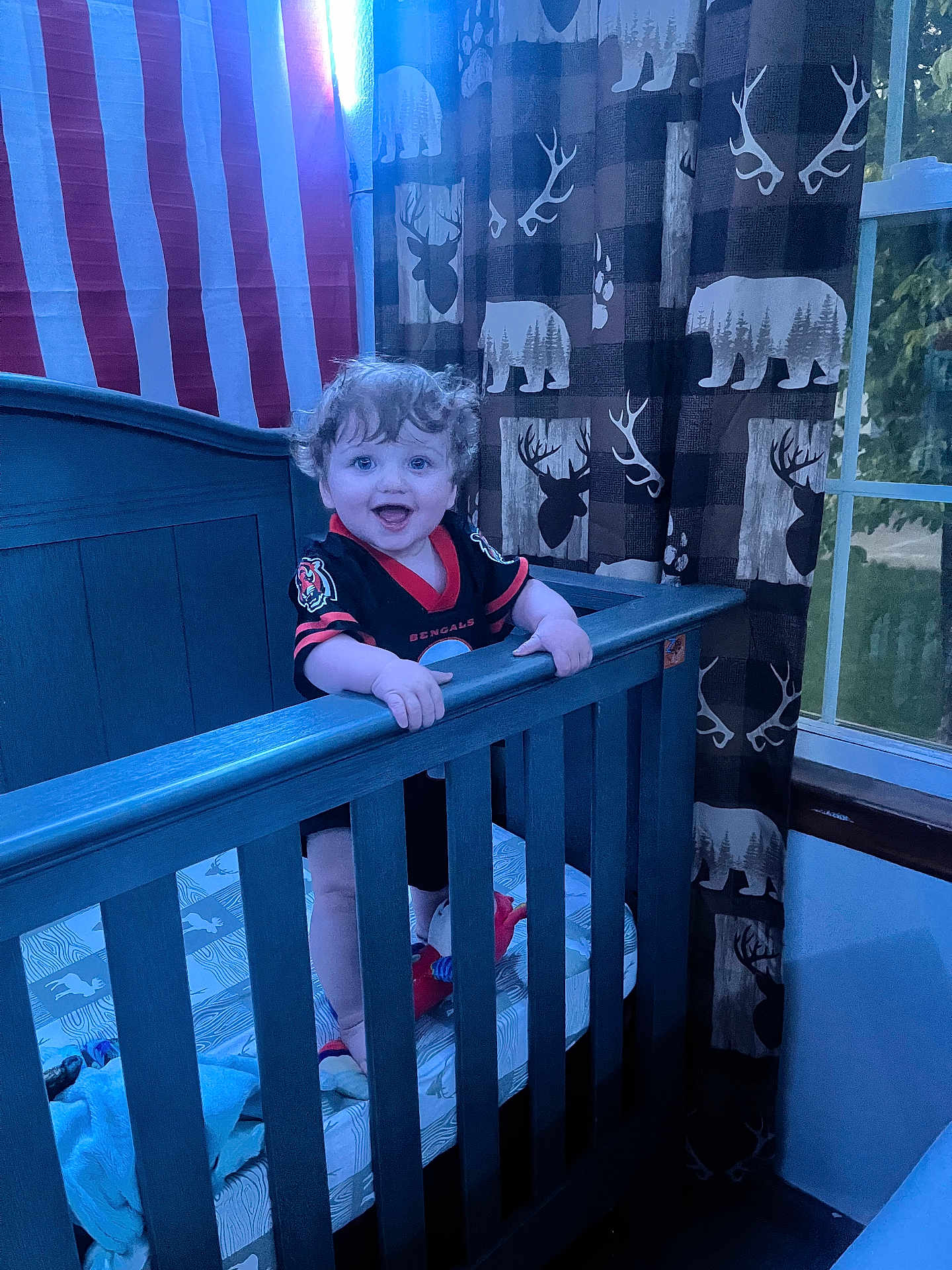 Parker is registered to the contest to win money with this photo: toddler, crib, child, curly_hair, smiling, indoor, curtains, window, bedroom, blue_crib, baby, clothing, patterned_curtains, toy, happy, person, wooden_furniture, standing, home, football_jersey