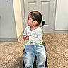 toddler, child, indoor, carpet, door, jeans, white_shoes, hair_tie, cardigan, stool, sitting, side_view, young_child, casual_clothing, small_furniture, wall, floor, portrait, person, cute