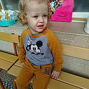 William a rejoint le concours — aidez-le/la à gagner de superbes lots ! toddler, child, bench, curly_hair, mickey_mouse, sweatshirt, pants, shoes, indoor, artwork, buttons, colorful, floor, portrait, expression, person, hair_tie, seat, wall, daycare