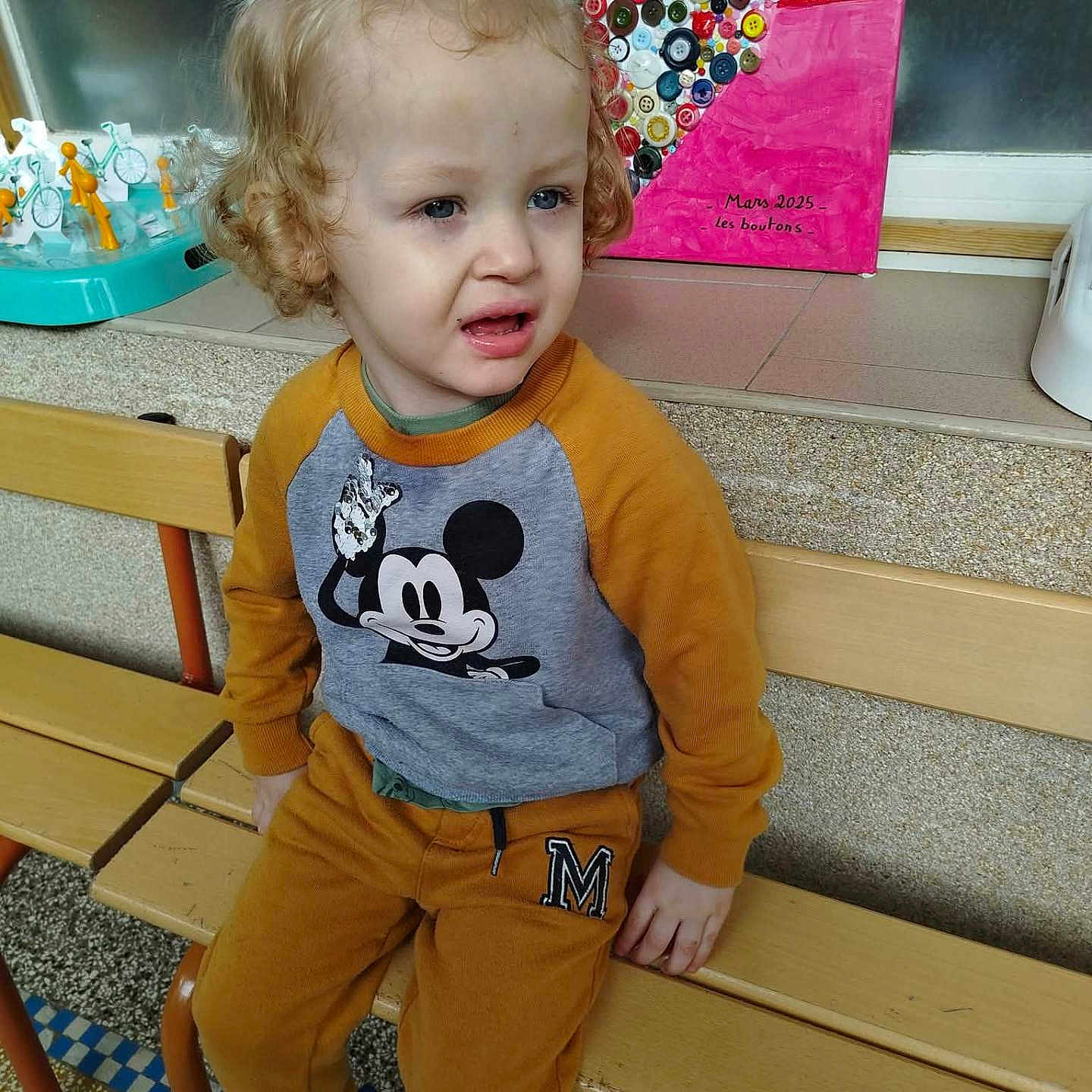 William a rejoint le concours — aidez-le/la à gagner de superbes lots ! toddler, child, bench, curly_hair, mickey_mouse, sweatshirt, pants, shoes, indoor, artwork, buttons, colorful, floor, portrait, expression, person, hair_tie, seat, wall, daycare