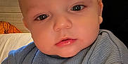 Brady is registered to the contest to win money with this photo: baby, infant, child, face, eyes, lips, mouth, nose, expression, portrait, close_up, blue_shirt, clothing, nike_logo, indoor, bed, pillow, curtain, skin, toddler