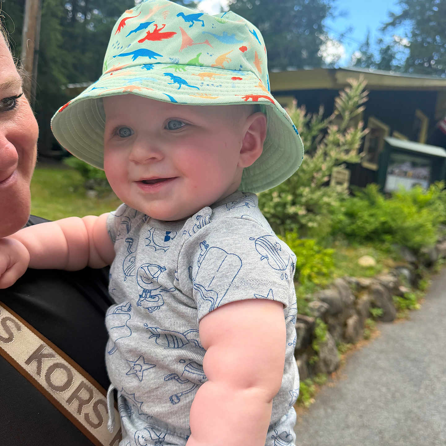 Sterlin is registered to the contest to win money with this photo: baby, child, smiling, hat, bucket_hat, dinosaur_pattern, outdoor, nature, greenery, trees, adult, holding, happy, face, cute, summer, sunny, cabin, pathway, clothing