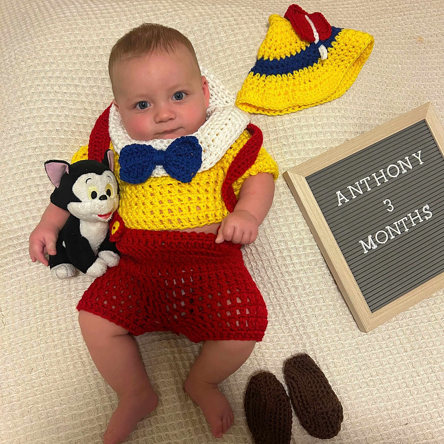 Anthony is registered to the contest to win money with this photo: baby, infant, child, portrait, smiling, crochet_outfit, yellow_hat, bow_tie, red_shorts, knitted_clothing, toy, stuffed_animal, letter_board, milestone, three_months, blanket, indoor, lying_down, colorful, family_photo