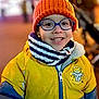 child, smiling, glasses, orange_hat, yellow_jacket, scarf, winter_clothing, outdoor, portrait, face, happy, cold_weather, nighttime, fashion, person, cute, colorful, casual, young, warm_clothing