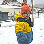 boots, child, cold_weather, daytime, fence, glasses, hat, house, jacket, outdoor, pants, person, playful, scarf, smile, snow, snowy_ground, standing, tree, winter_clothing