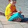 blue_pants, boy, bubble_wand, casual_clothing, child, daytime, footwear, glasses, happy, orange_shirt, outdoor, pavement, person, playful, relaxed, sitting, smiling, sunny, urban, young
