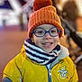 astronaut_patch, child, closeup, cold_weather, cute, face, fashion, glasses, happy, kid, orange_hat, outdoor, pom_pom, portrait, scarf, smiling, striped_scarf, warm_clothes, winter_clothing, yellow_jacket