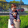 child, boy, glasses, outdoor, grass, toy, sneakers, fence, sunlight, smile, greenery, playful, casual_clothing, daylight, young, person, nature, happy, sitting, park