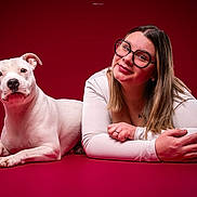 Joy participe au concours pour gagner de l'argent avec cette photo : dog, woman, portrait, studio, red_background, white_dog, glasses, smiling, lying_down, pet, companion, photography, indoors, dramatic_lighting, closeup, human, animal, white_shirt, long_hair, tattoo