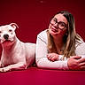 dog, woman, portrait, studio, red_background, white_dog, glasses, smiling, lying_down, pet, companion, photography, indoors, dramatic_lighting, closeup, human, animal, white_shirt, long_hair, tattoo