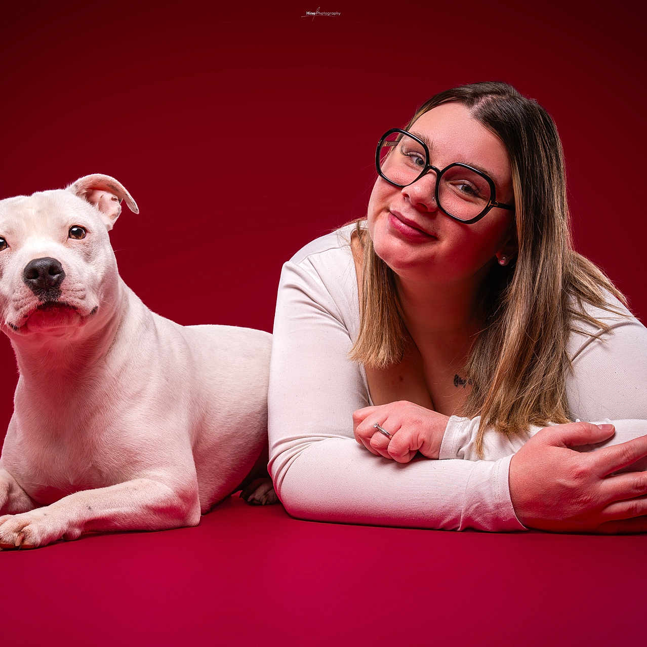 Joy participe au concours pour gagner de l'argent avec cette photo : dog, woman, portrait, studio, red_background, white_dog, glasses, smiling, lying_down, pet, companion, photography, indoors, dramatic_lighting, closeup, human, animal, white_shirt, long_hair, tattoo